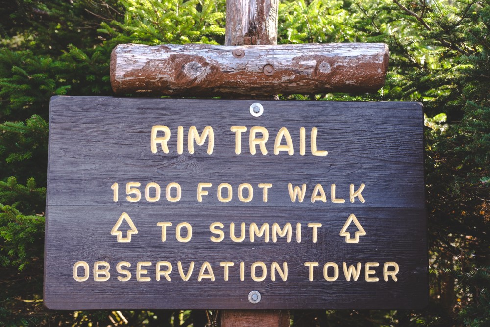 A wooden sign with carved and painted text that reads "RIM TRAIL 1500 FOOT WALK TO SUMMIT OBSERVATION TOWER" with arrow symbols pointing upwards. The sign is mounted on rustic wooden posts.