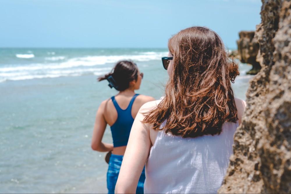 Two women at the beach, one with brown hair wearing a white top looking towards the sea, the other wearing a blue top and black sunglasses, facing away from the camera towards the ocean. Waves and a rocky formation are seen in the background