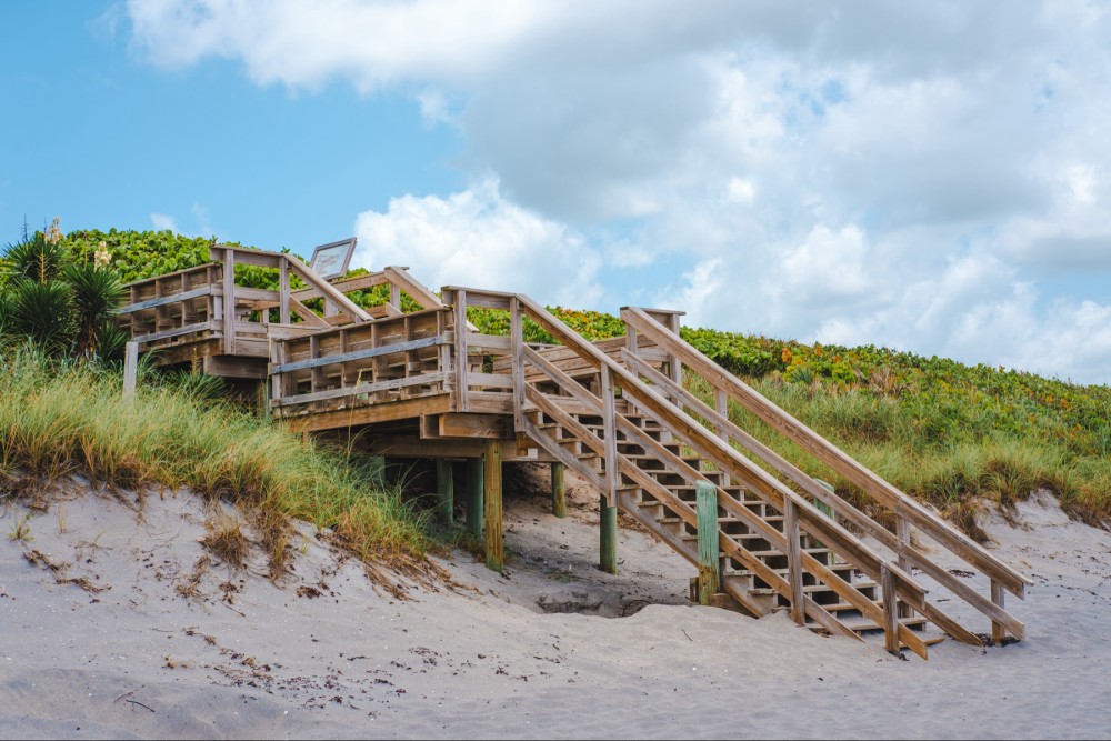 A wooden staircase over a sandy dune leading up to a beach access point with vegetation on either side under a partly cloudy sky.