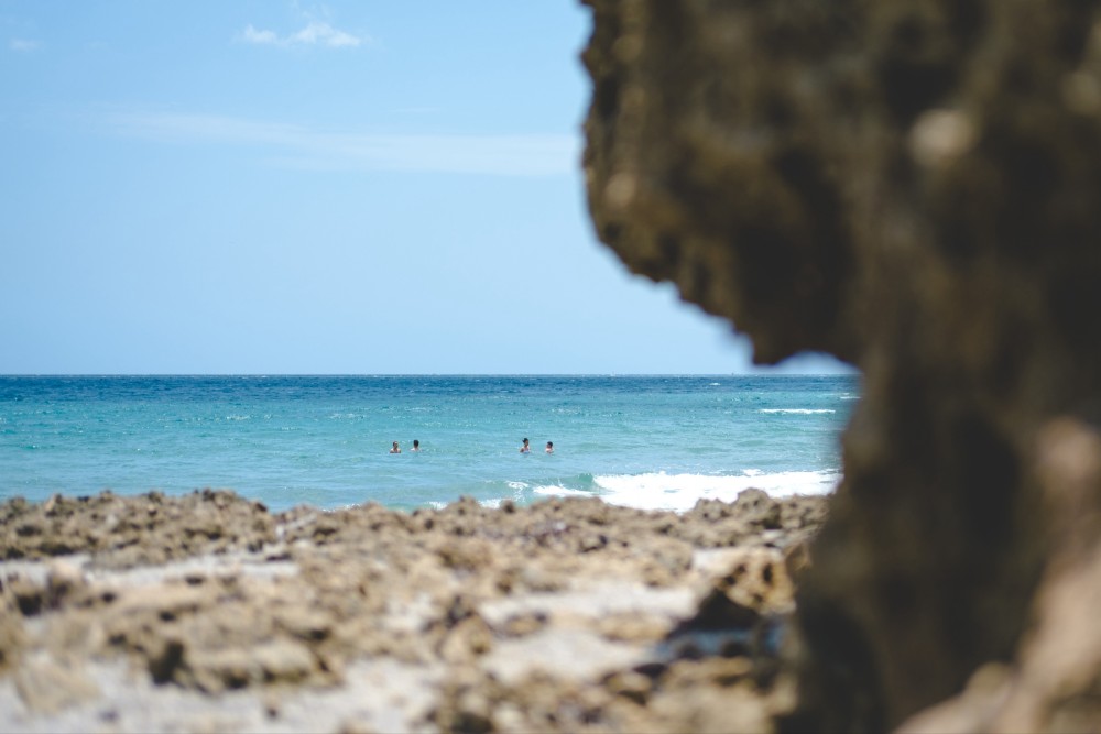 Beach scene with people swimming in the sea, viewed through a natural rocky frame. Clear blue sky above gentle turquoise waves.