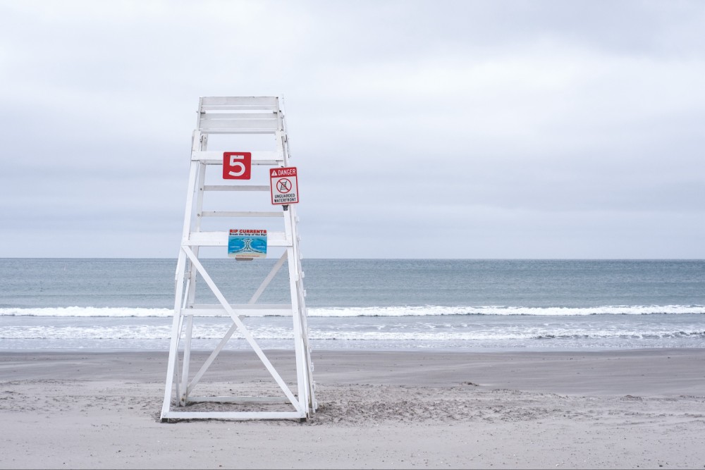 A lifeguard tower with the number 5 and warning signs about an unguarded waterfront and rip currents on a sandy beach with the ocean in the background on an overcast day.