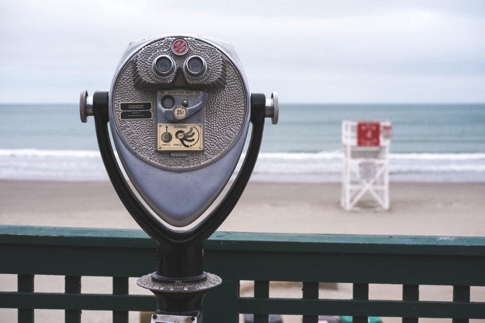 Coin-operated viewing binoculars on a railing overlooking a beach with a lifeguard tower in the distance on an overcast day.