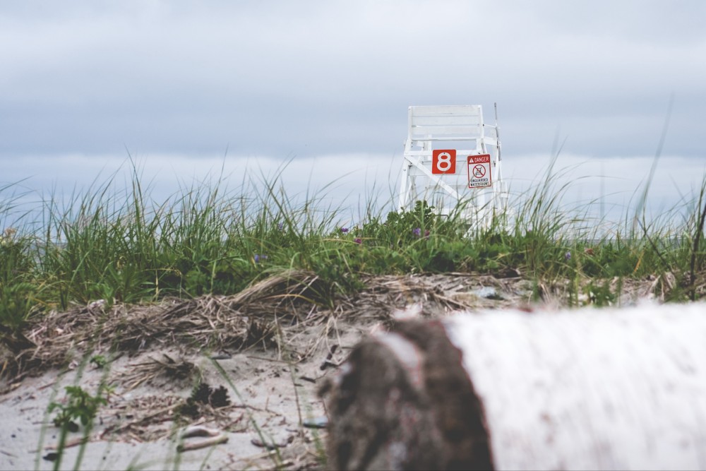 A lifeguard chair with a number "8" stands beyond a grassy sand dune on a cloudy day, with a warning sign indicating no lifeguard on duty.