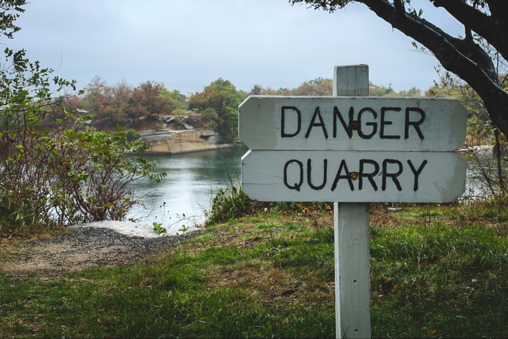 A warning sign that reads "DANGER QUARRY" near a body of water with trees and overcast skies in the background.