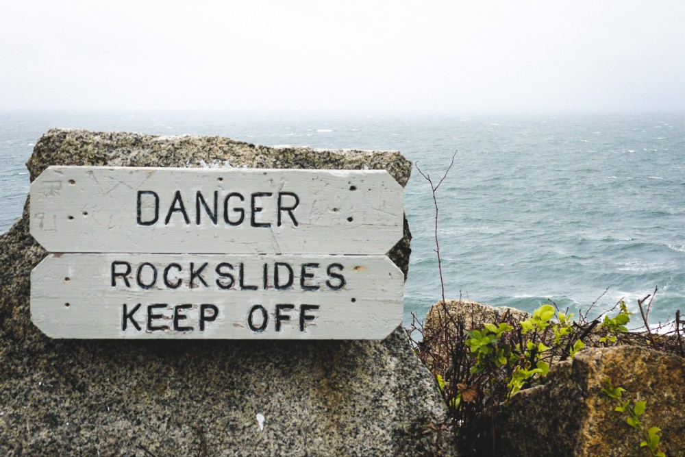 A weathered white warning sign with black lettering that reads "DANGER ROCKSLIDES KEEP OFF" alongside a rocky coastline with a rough sea in the background.