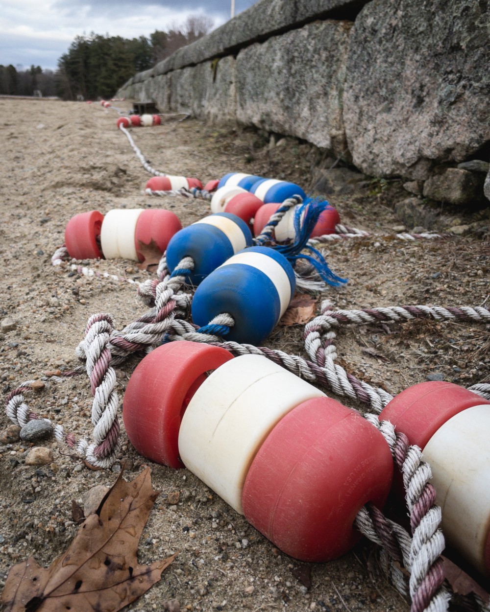 A series of colorful buoy floats attached to ropes, lined up along a sandy ground near a stone wall, under a cloudy sky.