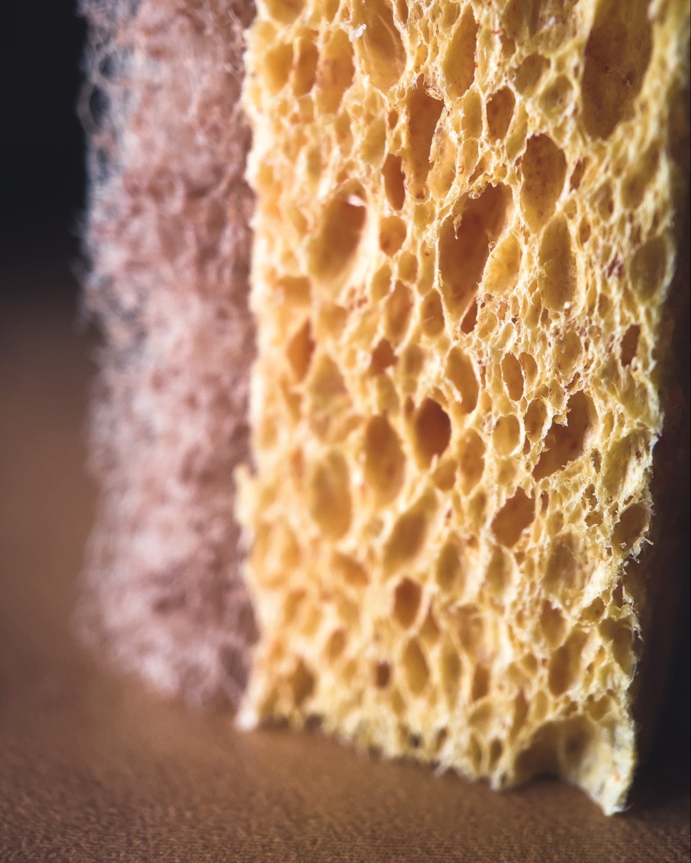 Macro photo of the bottom corner of a yellow cleaning sponge with a brown textured scrubbing pad on top of a warm brown wood surface.