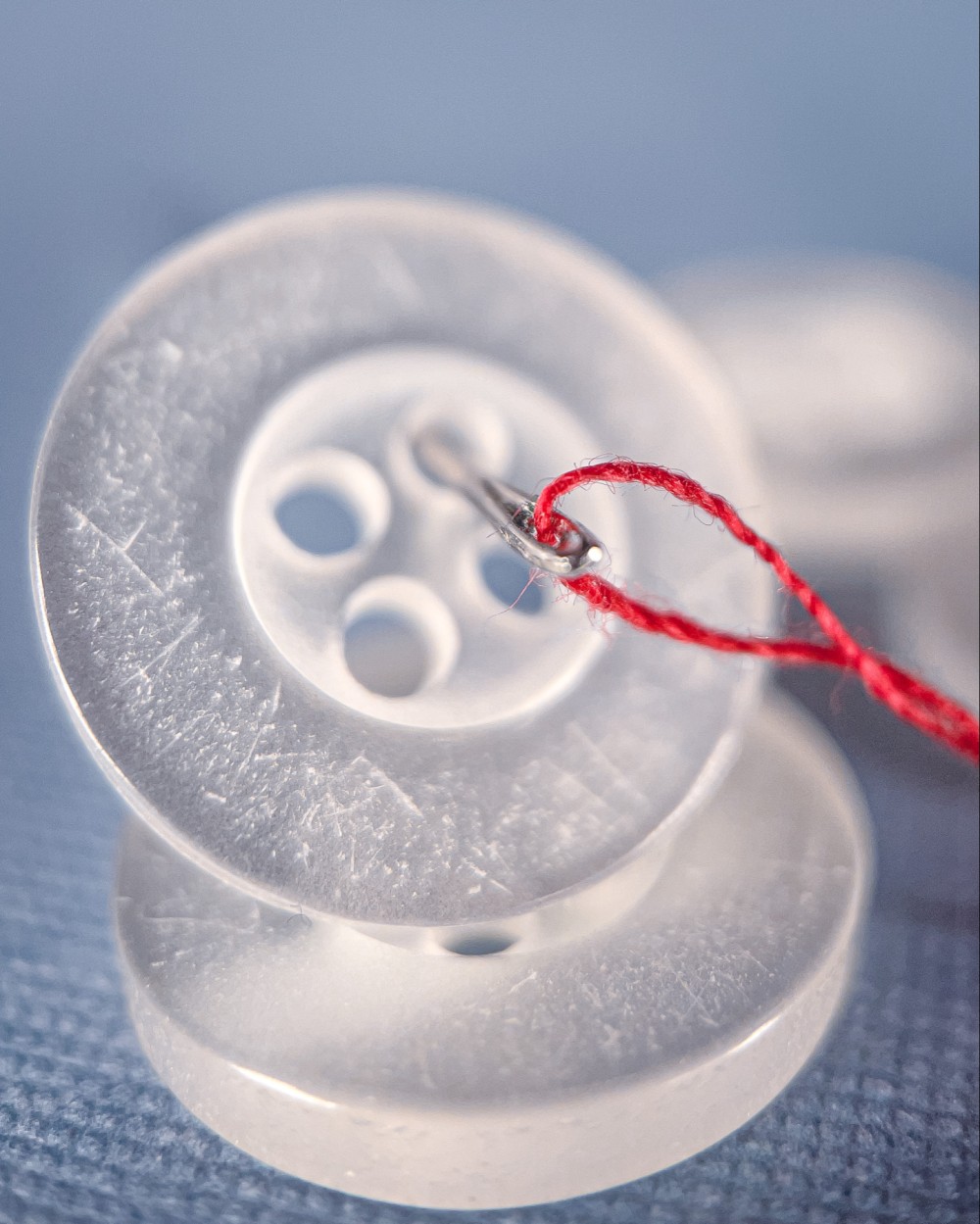 Macro photo of two translucent white clothes buttons stacked on top of each other with more blurred buttons behind them on a blue background. The top button in the stack has a needle attached to a red piece of thread running through it.