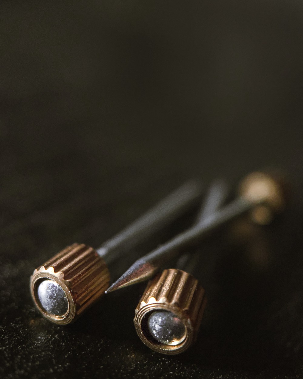 Macro photo of three silver nails with textured brass cylinders just under each of their heads. Nails are on top of and in front of a black background.