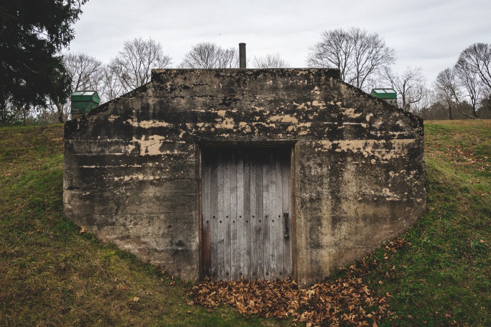 A small, weathered concrete bunker with a wooden door set into a grassy hillside, surrounded by leafless trees in the background.