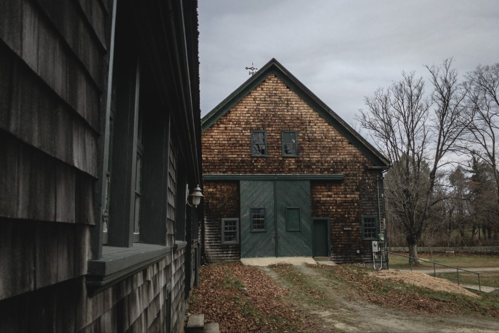 A rustic building with wooden shingles and a large green double door. The structure is flanked by another building on the left, with leaf-strewn ground leading up to the entrance.