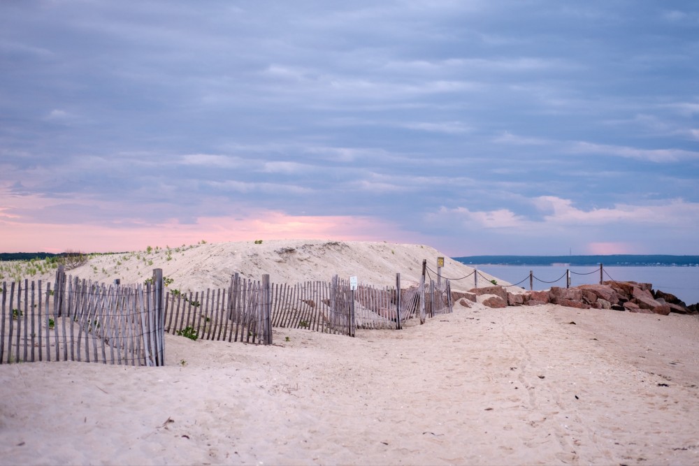 A sandy beach at sunrise with a wooden fence, dunes, and plants, against a backdrop of a sunrise sky with shades of pink and blue.