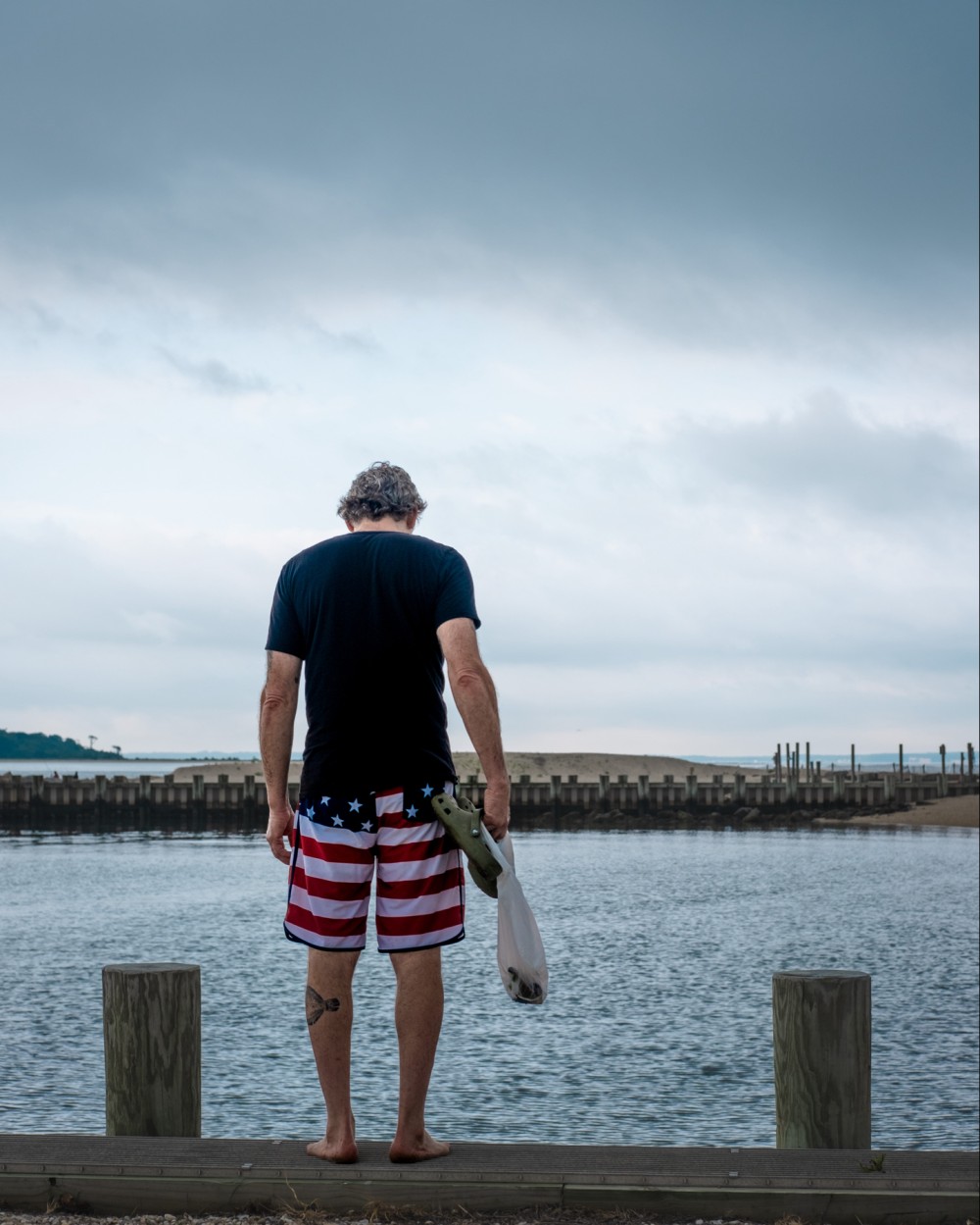 A man standing on a dock, facing the ocean, wearing American flag shorts and holding a pair of sandals. The sky is overcast.