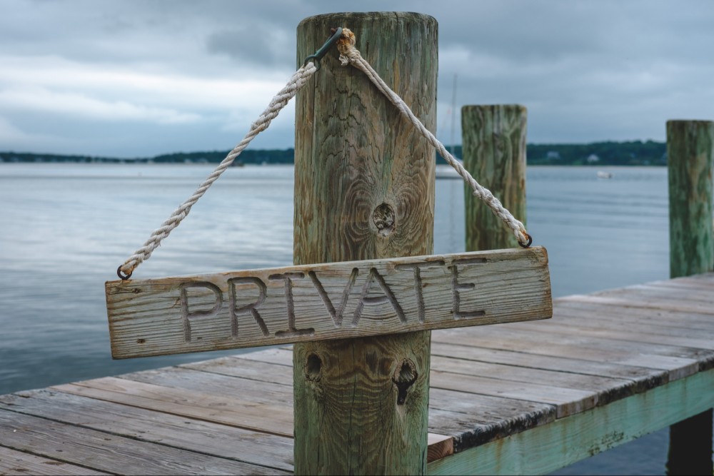 A wooden sign with the word "PRIVATE" hangs by a rope on a wooden post at a dock with a tranquil water backdrop.