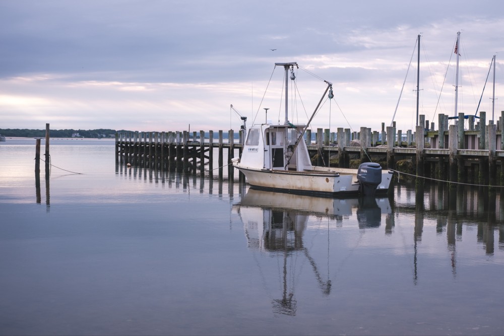 A tranquil harbor with a moored fishing boat, a wooden pier with vertical pylons, and still water reflecting the sky.