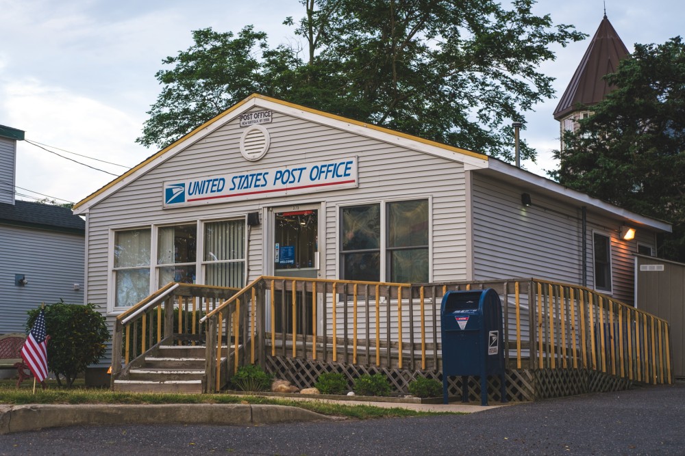 A small United States Post Office building with a wooden ramp entrance, an American flag, and a blue USPS collection mailbox outside.
