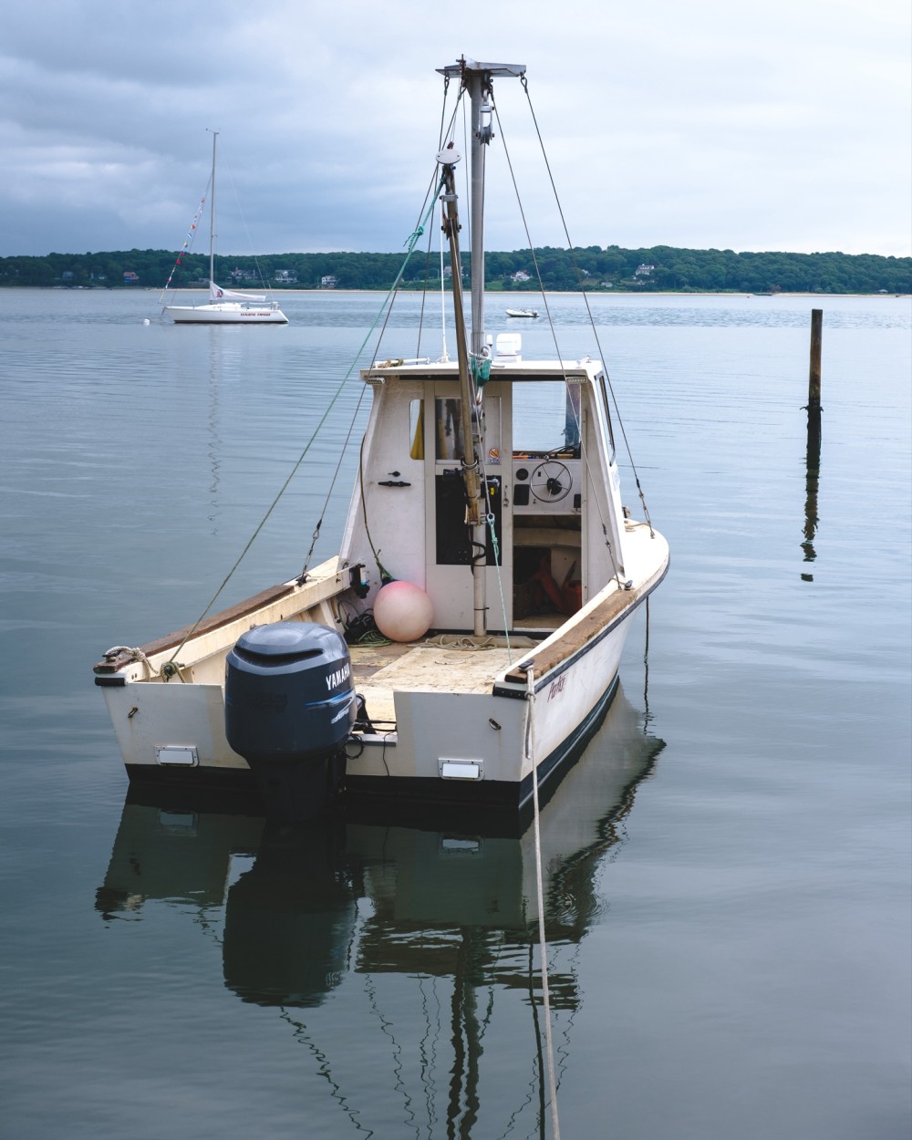 A small fishing boat moored in calm waters with a mast and Yamaha outboard motor, with another sailboat and shoreline in the distance.
