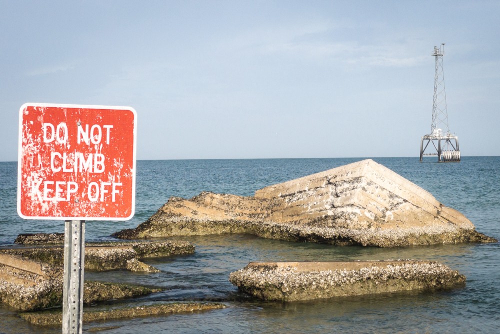 A weathered red sign with the words "DO NOT CLIMB KEEP OFF" stands by the sea, with concrete structures covered in barnacles in the water and a distant metal tower on stilts visible in the background.