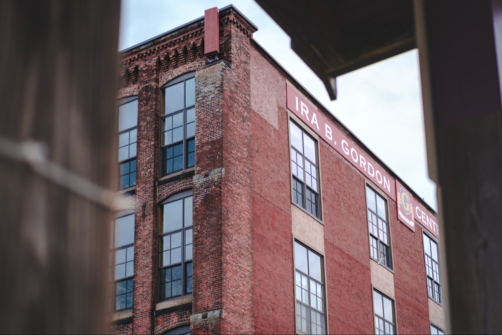 An upward shot of an industrial brick building with large windows and a sign that reads "IRA B. GORDON CENTER” framed through a wooden structure