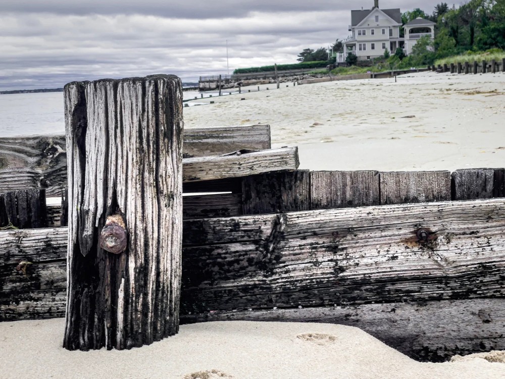 Close up photo of a weathered wooden fence along a beach. In the background is a stormy sky and far away house surrounded by sand and grass.