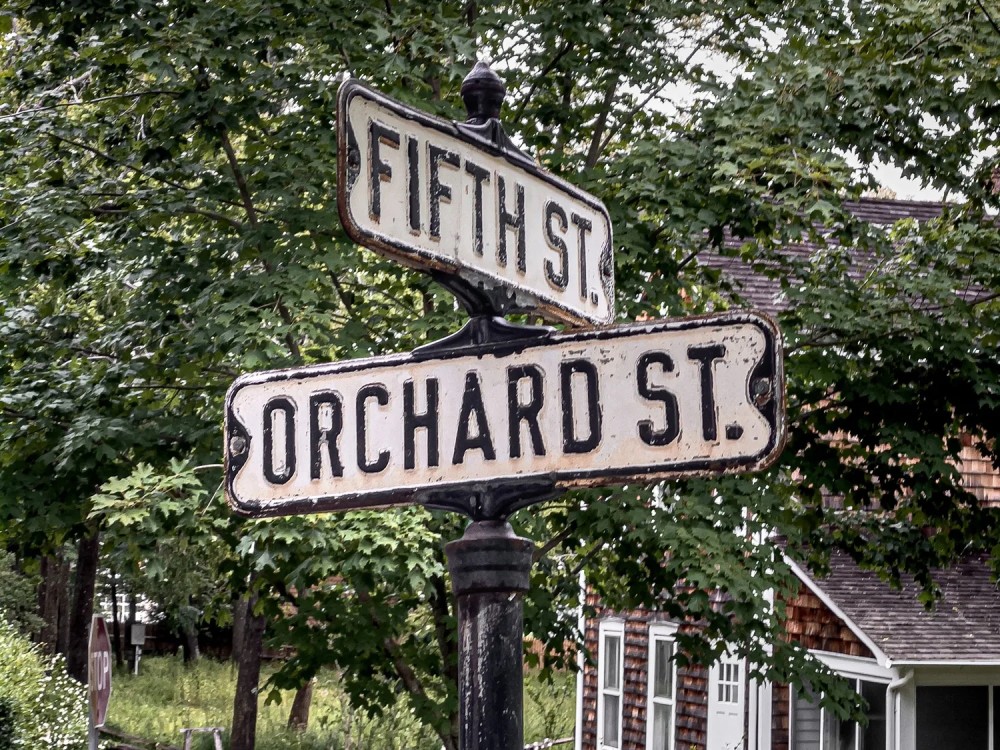 Close up of two intersected street signs that read “fifth st” and “orchard st”. Signs are white with black text mounted on a black pole in front of trees and a brown house.