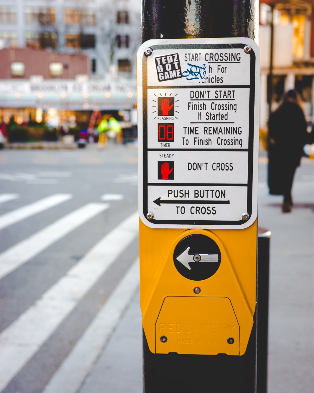 A pedestrian crosswalk signal button with instructions. In the background a person walks along the sidewalk