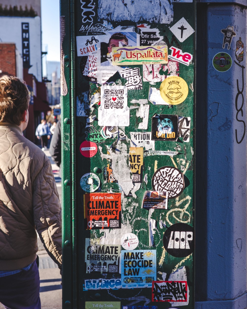 A green box connected to a traffic pole mostly filled with various stickers. A person is partially visible walking on a nearby crosswalk
