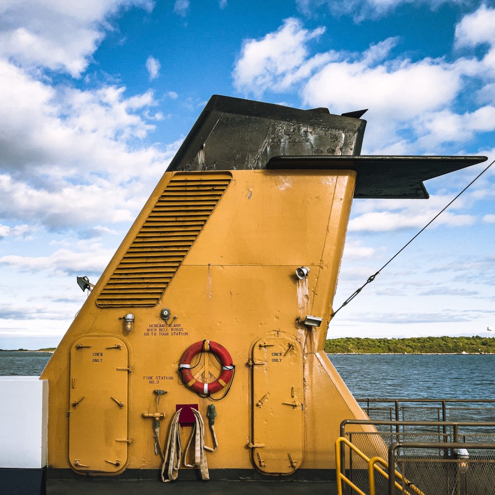 Yellow ferry boat funnel with a red lifebuoy and "Crew Only" sign, against a backdrop of water on a sunny day with fluffy white clouds in the sky.