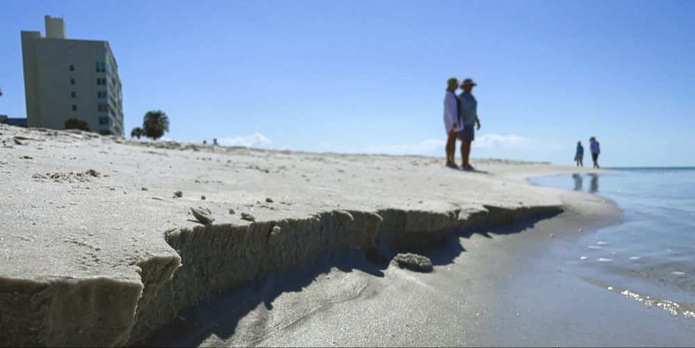 Close up photo of a self of sand meeting the water on a beach in front of a bright blue sky. In the background, there is a single building behind two people looking into the ocean and two walking along the beach.