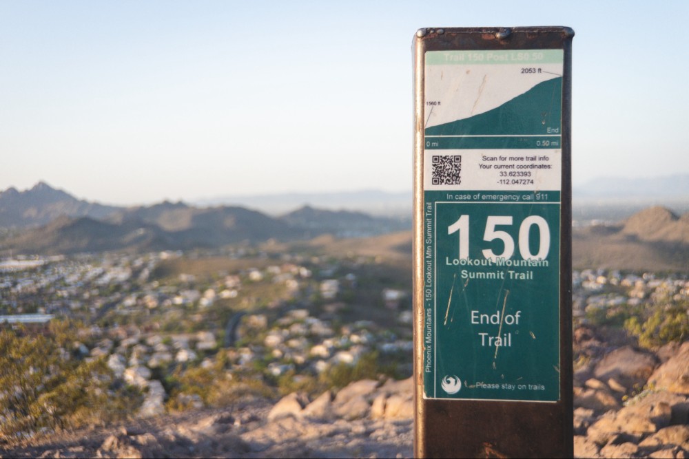 A trailhead sign for Lookout Mountain Summit Trail with the number 150, marking the end of the trail. In the background, a blurred view of a mountainous landscape and cityscape is visible under a clear sky.