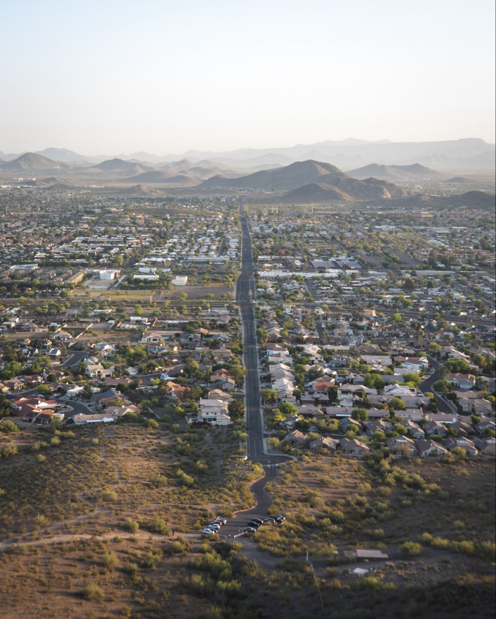 Aerial view of a suburban area with a long straight road extending towards the horizon, flanked by houses, with mountainous terrain in the distance under a soft-lit sky.