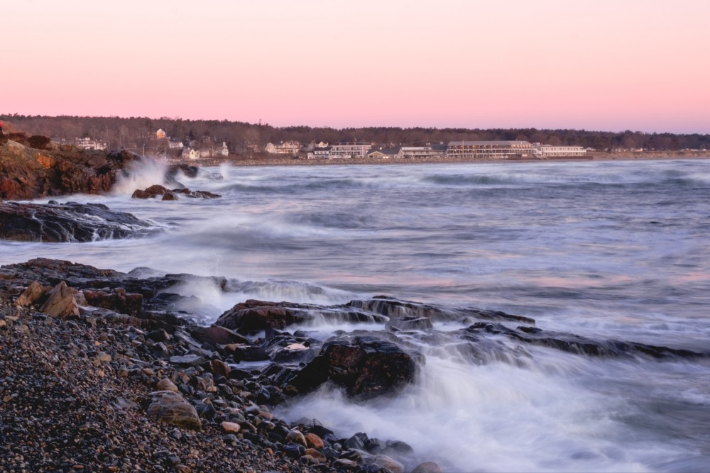 Seascape at sunrise with waves crashing against rocky shoreline, pebbly beach in foreground, and large waterfront buildings in the distance under a pink-hued sky.