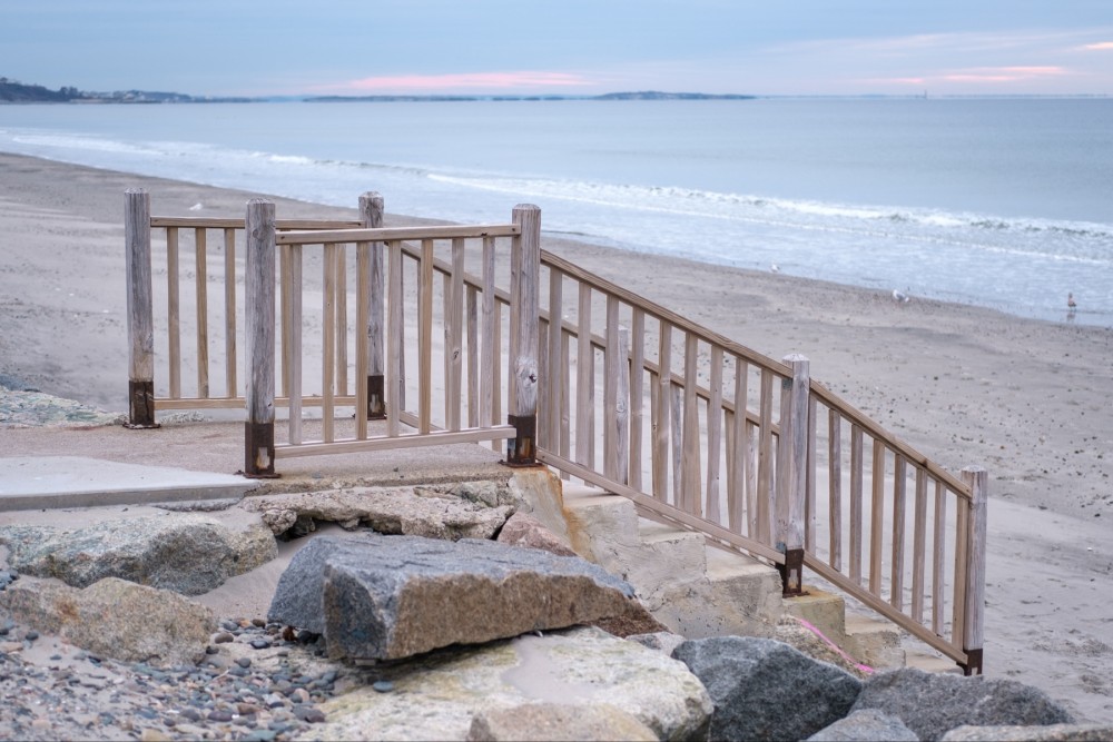 Wooden stairs leading down to a sandy beach with rocks in the foreground and the ocean in the background a pale sunrise.