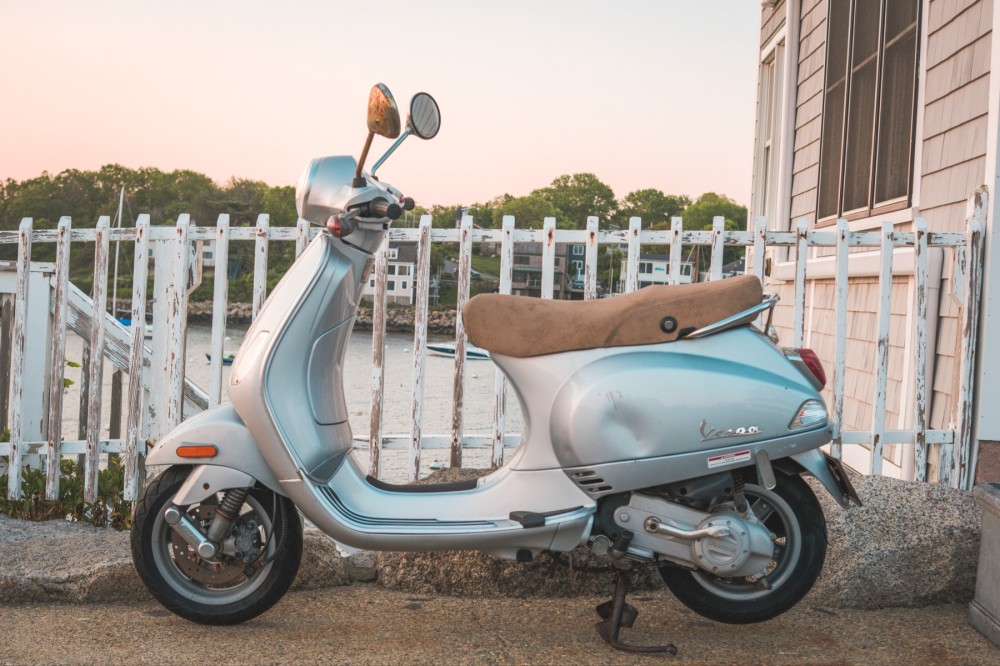 A silver Vespa scooter parked in front of a white picket fence, with a coastal background at sunrise.