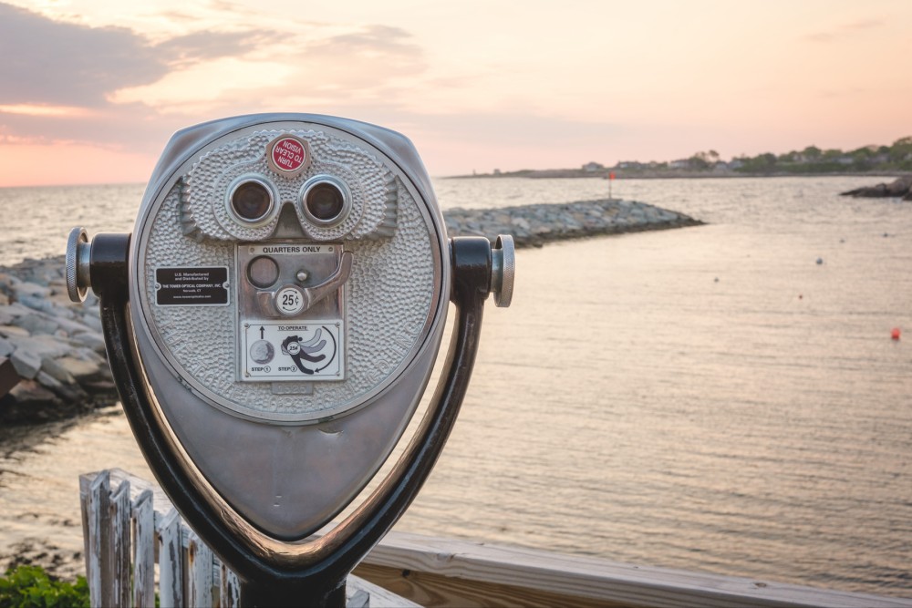 Coin-operated binoculars overlooking the ocean at sunrise with rocks and a breakwater in the background.