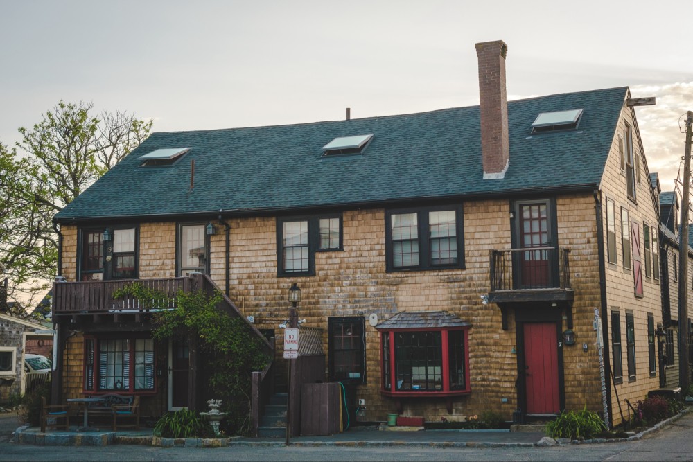 A two-story shingled building with a green roof, featuring multiple windows and red doors, a chimney, and a "No Parking" sign, set against a morning sky.