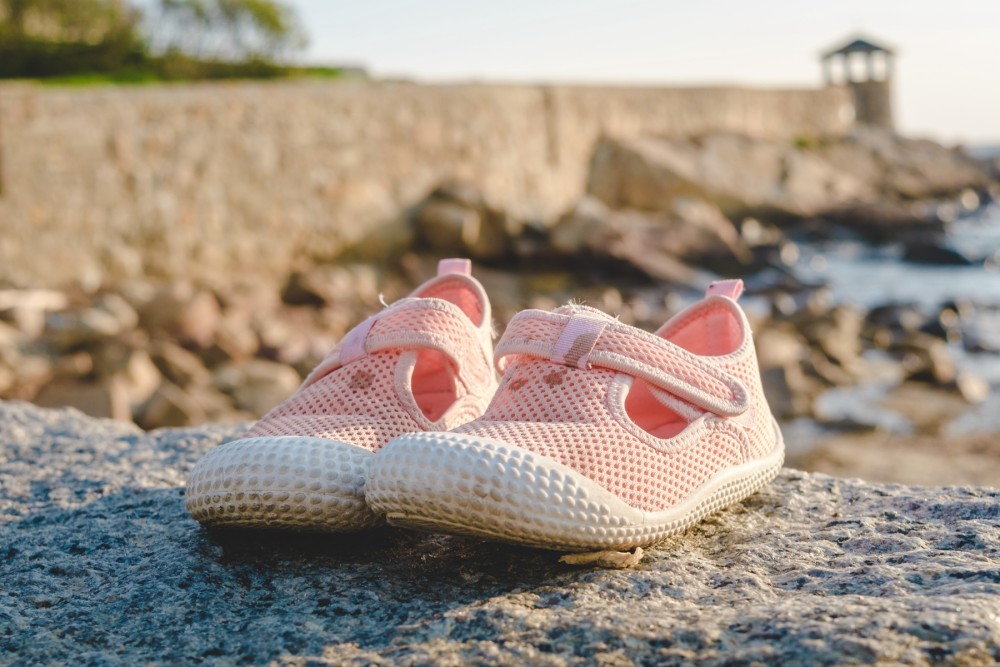 A pair of pink children's shoes on a rocky surface with a blurred background of a stone wall and water.