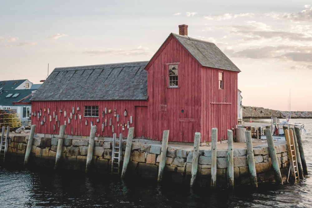 A red wooden building (Motif #1) on a stone pier by the water, with pilings and ladders leading to the sea, at sunrise. A boat is docked nearby.